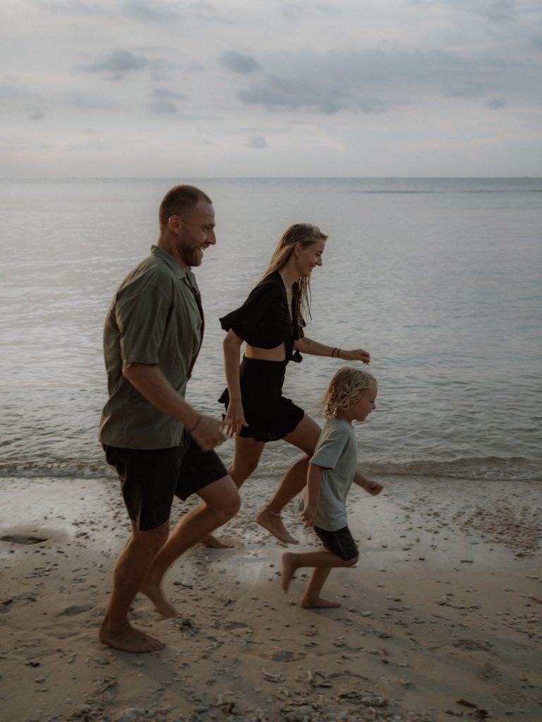 Familie läuft barfuß am Strand entlang, Wolken am Himmel über dem Wasser.