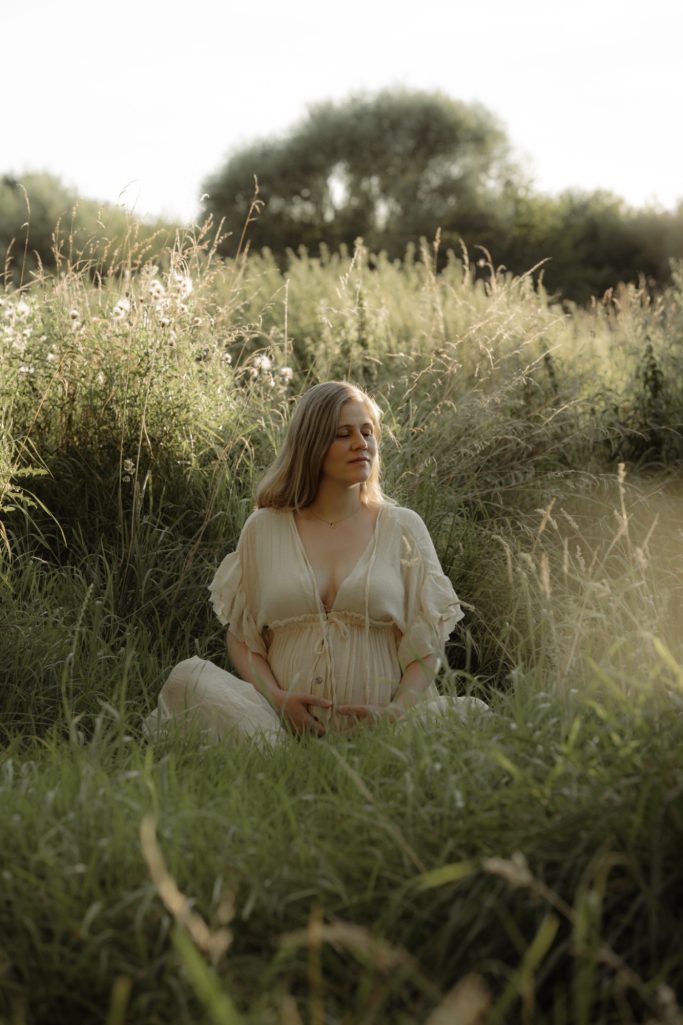 Schwangere Frau in weißem Kleid sitzt im hohen Gras unter natürlichem Licht.