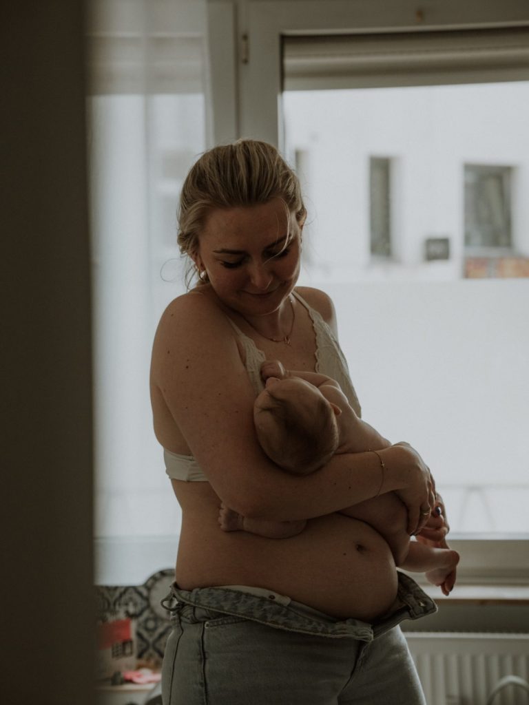 Eine Mutter stillt ihr Baby in einem hellen Raum mit großen Fenstern.
